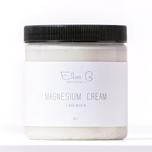 Puede incluir: Un tarro transparente con tapa negra, lleno de crema blanca. Una etiqueta blanca muestra el texto "Ellie B Naturals MAGNESIUM CREAM LAVENDER 8oz". El tarro está sobre un fondo blanco.