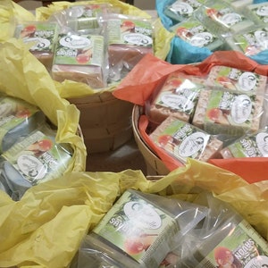 May include: Four baskets filled with wrapped bars of soap. The baskets are lined with yellow, orange, and blue tissue paper. The soap bars are wrapped in clear plastic and have a green and white label with the text "Norms Savon" on them.