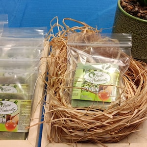 May include: A close-up of a wooden crate filled with small clear plastic bags containing green soap bars. The soap bars have a label that reads "Juste Naturelle" and "Savon". The crate is surrounded by straw-like material. There are also several small cards with the text "Savon" on them.