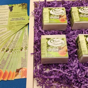 Peut inclure: Quatre savons naturels dans une boîte cadeau avec du papier de soie violet. Le savon est étiqueté "Juste Naturelle Savon Miel et Olive".