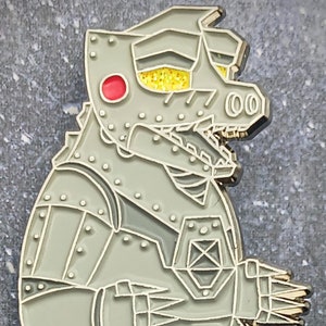 May include: A gray metal pin depicting a robotic dinosaur with a red eye, yellow glitter in the eye area, and silver accents. The pin is in a side profile, showcasing its detailed mechanical design and rivets.