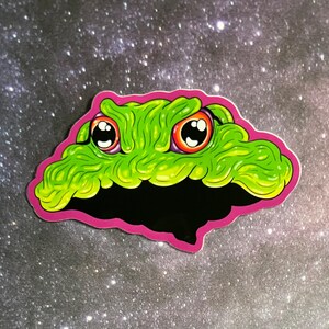 May include: A green, gooey, cartoon monster with large, red eyes and a pink outline. The monster has a menacing expression and appears to be floating in space.
