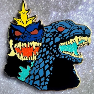 May include: A metal enamel pin featuring two stylized monster heads. The left head is dark blue with a yellow crown, red eyes, and a glittery orange mouth. The right head is dark blue with a textured scale pattern, red eyes, and a glittery teal mouth.