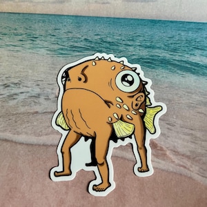 May include: A cartoon sticker of a brown pufferfish with a human-like body and legs walking on a sandy beach. The fish has a large eye and a spiky, textured body.