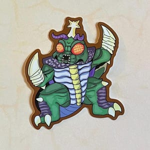 May include: A green and purple monster with a star on its head, wearing a purple and white striped shirt. The monster has large red eyes and a toothy grin. The monster is standing on a brown background.