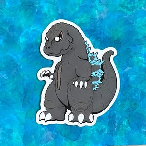 May include: A gray Godzilla sticker with a white border, featuring light blue dorsal fins and tail spikes. The sticker is set against a blue and teal watercolor background. The cartoon-style design has a stern expression.