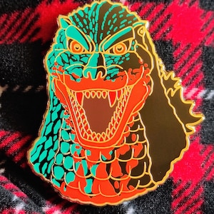 May include: A Godzilla enamel pin with a gold outline. The pin features a detailed design of Godzilla's head, with teal, orange, and black colors. The pin is set against a red and black plaid background.