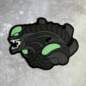 May include: Black and green alien creature sticker with a menacing expression. The alien has a detailed, segmented body and glowing green eyes.