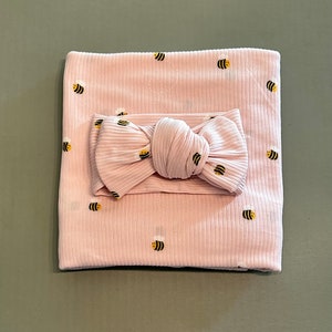 May include: A light pink baby blanket and matching headband set. The blanket and headband are ribbed and feature a repeating pattern of embroidered yellow and black bees. The headband is tied in a bow.