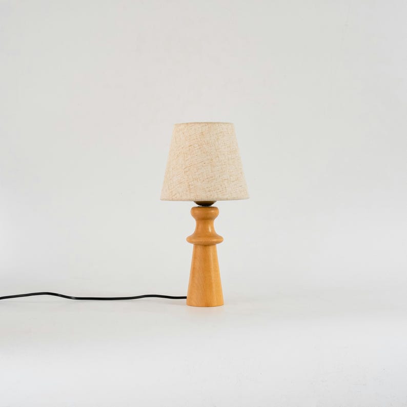 May include: A wooden table lamp with a beige linen lampshade. The lamp has a simple, cylindrical design with a slightly flared base.