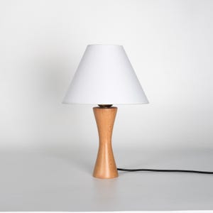 May include: A small, wooden table lamp with a white lampshade. The lamp has a light brown, hourglass-shaped base and a white conical shade. A black power cord extends from the base.