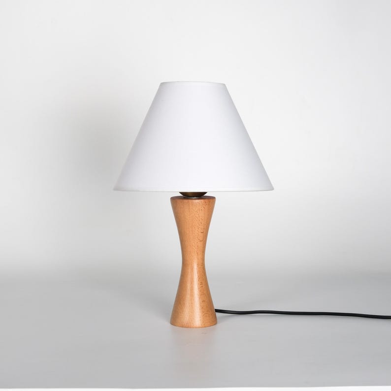 May include: A small, wooden table lamp with a white lampshade. The lamp has a light brown, hourglass-shaped base and a white conical shade. A black power cord extends from the base.