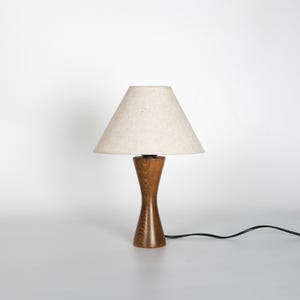 May include: A wooden table lamp with a beige lampshade. The lamp has a hourglass-shaped base made of dark brown wood. A black power cord extends from the base. The lampshade is a light beige color.