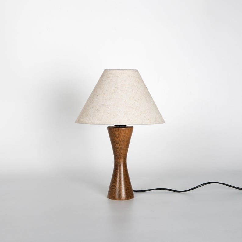 May include: A wooden table lamp with a beige lampshade. The lamp has a hourglass-shaped base made of dark brown wood. A black power cord extends from the base. The lampshade is a light beige color.