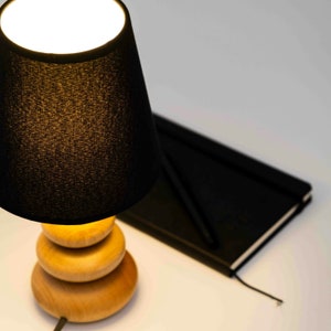 Pode incluir: Um abajur preto com uma base de madeira. A base &eacute; feita de tr&ecirc;s c&iacute;rculos empilhados. A l&acirc;mpada est&aacute; ligada e iluminando o quarto.