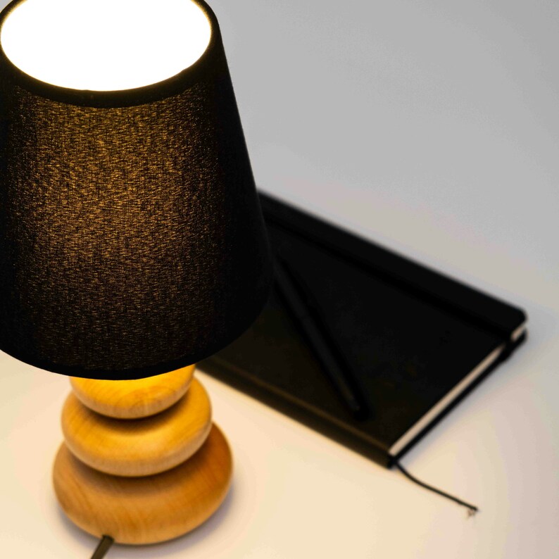 Pode incluir: Um abajur preto com uma base de madeira. A base &eacute; feita de tr&ecirc;s c&iacute;rculos empilhados. A l&acirc;mpada est&aacute; ligada e iluminando o quarto.