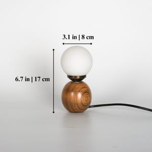 May include: A contemporary table lamp featuring a wooden base and a white, spherical shade. The lamp measures 17 cm in height and 8 cm in width. The base has a wood grain design and a brass-toned detail.
