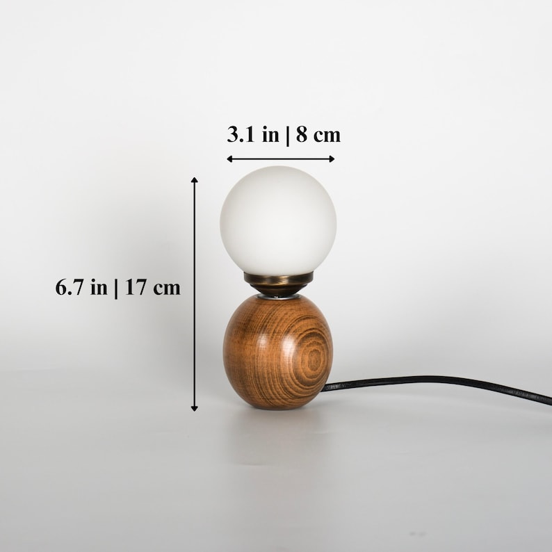 May include: A contemporary table lamp featuring a wooden base and a white, spherical shade. The lamp measures 17 cm in height and 8 cm in width. The base has a wood grain design and a brass-toned detail.
