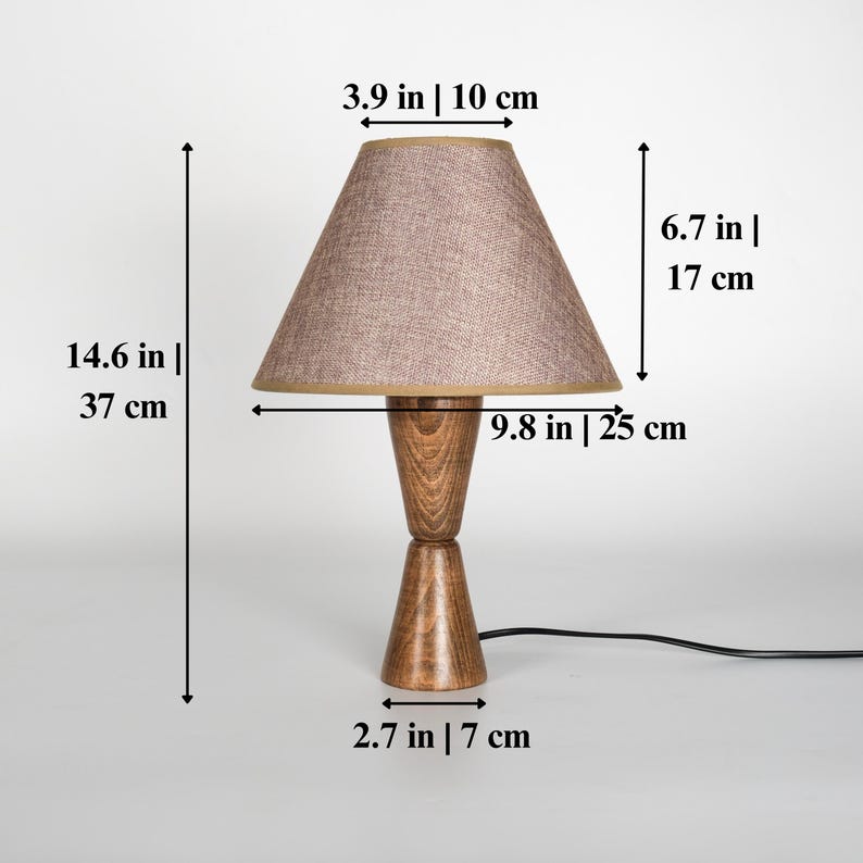 May include: A wooden table lamp with a beige fabric shade. The lamp base is made of two stacked, cone-shaped pieces of wood. The lamp is 14.6 inches (37 cm) tall, with a shade that is 9.8 inches (25 cm) wide.