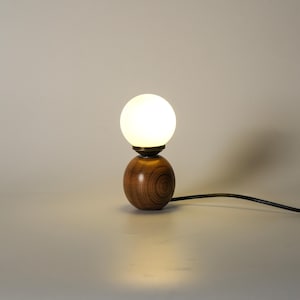 May include: A small wooden table lamp with a spherical base and a round, frosted glass bulb. The lamp emits a warm glow and has a dark power cord. The wooden base displays a visible wood grain pattern.