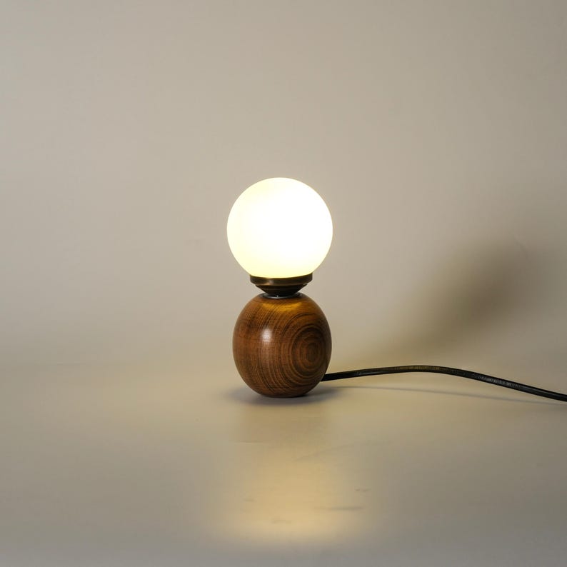 May include: A small wooden table lamp with a spherical base and a round, frosted glass bulb. The lamp emits a warm glow and has a dark power cord. The wooden base displays a visible wood grain pattern.