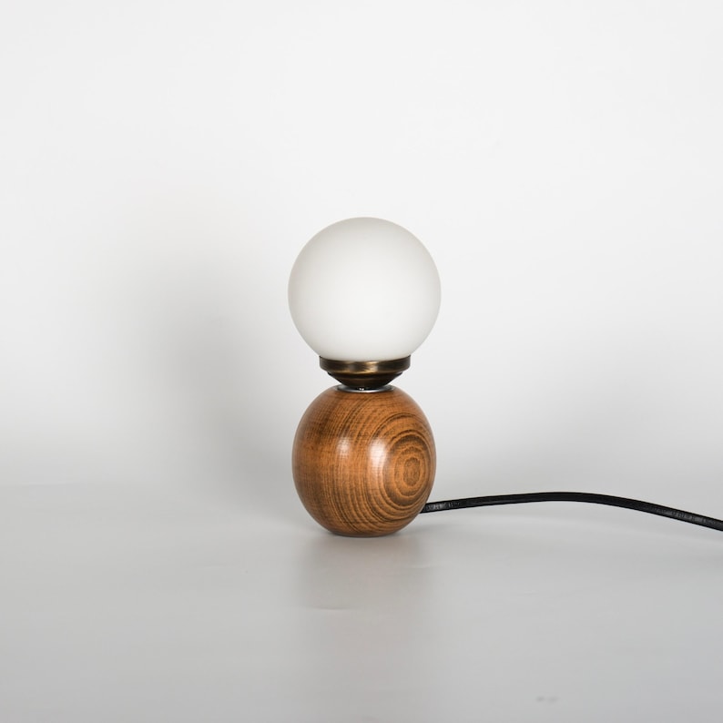 May include: A small table lamp featuring a wooden base and a white, globe-shaped light. The base is a warm brown with a wood grain pattern. The lamp is connected to a black power cord.
