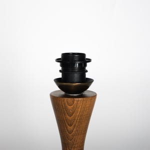 May include: A wooden lamp base with a black lamp socket and a bronze-colored metal accent. The base is a warm brown color with a conical shape. The lamp socket is made of black plastic.