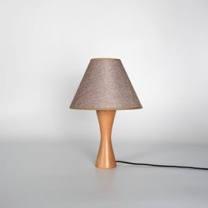 May include: A small, wooden table lamp with a light brown, conical lampshade. The base is a light-colored wood with a curved hourglass shape. A black power cord extends from the base.