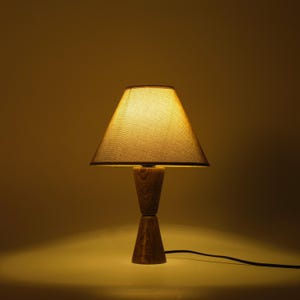 May include: A wooden table lamp with a beige lampshade. The lamp has a unique hourglass-shaped base and a conical shade. The lamp is lit, casting a warm glow. The cord is black.