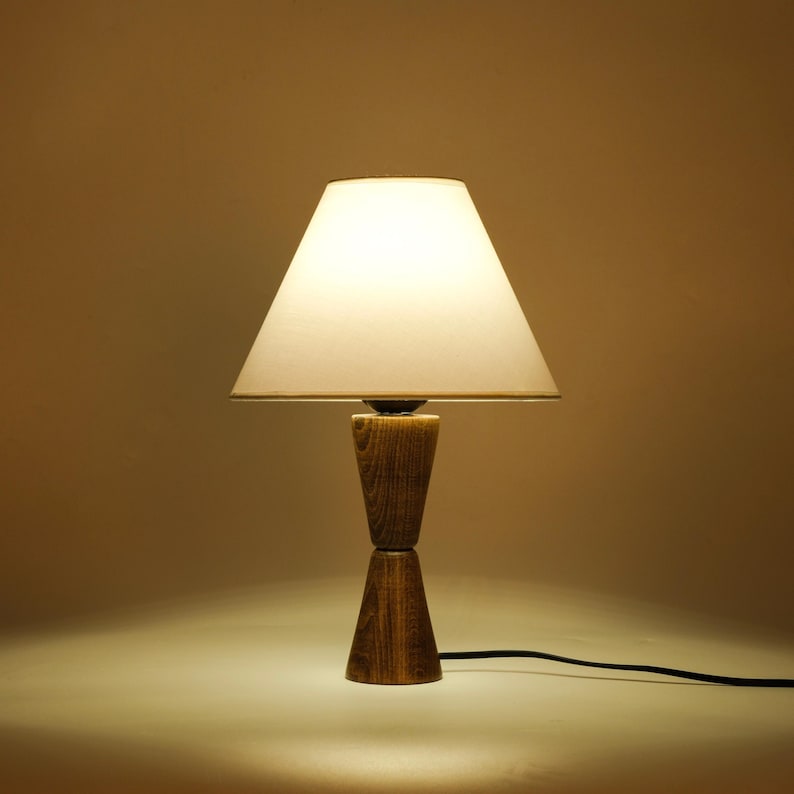 May include: A wooden table lamp with a white lampshade. The lamp has a unique hourglass-shaped base made of wood. The lampshade is a light cream color, and the lamp is plugged into a black cord.