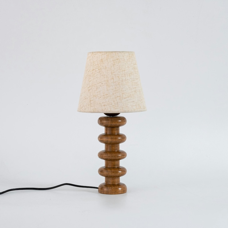 May include: A wooden table lamp with a natural wood finish and a beige fabric lampshade. The lamp base is made of stacked, rounded pieces of wood.