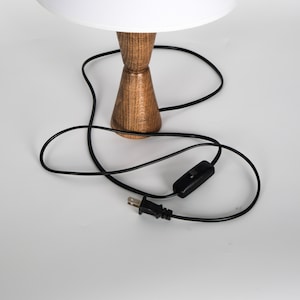 May include: A wooden table lamp with a white lampshade. The lamp has a brown, hourglass-shaped base and a black power cord with an on/off switch and a two-prong plug.