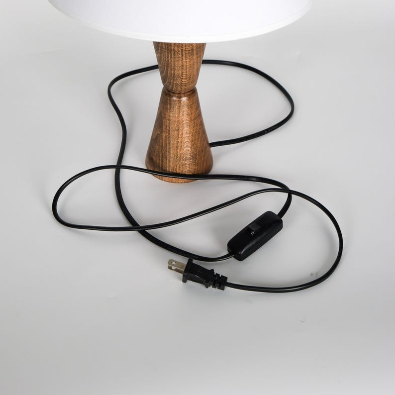 May include: A wooden table lamp with a white lampshade. The lamp has a brown, hourglass-shaped base and a black power cord with an on/off switch and a two-prong plug.