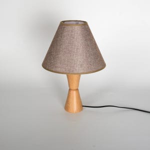 May include: A small table lamp with a light brown fabric shade and a wooden base. The shade is cone-shaped with a gold trim. The base is made of light-colored wood and has a unique hourglass shape. A black power cord extends from the base.