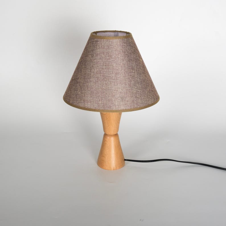 May include: A small table lamp with a light brown fabric shade and a wooden base. The shade is cone-shaped with a gold trim. The base is made of light-colored wood and has a unique hourglass shape. A black power cord extends from the base.