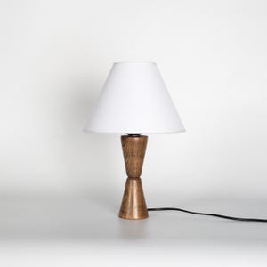 May include: A wooden table lamp with a white lampshade. The lamp has a two-part, hourglass-shaped base made of light brown wood. A black power cord extends from the base.