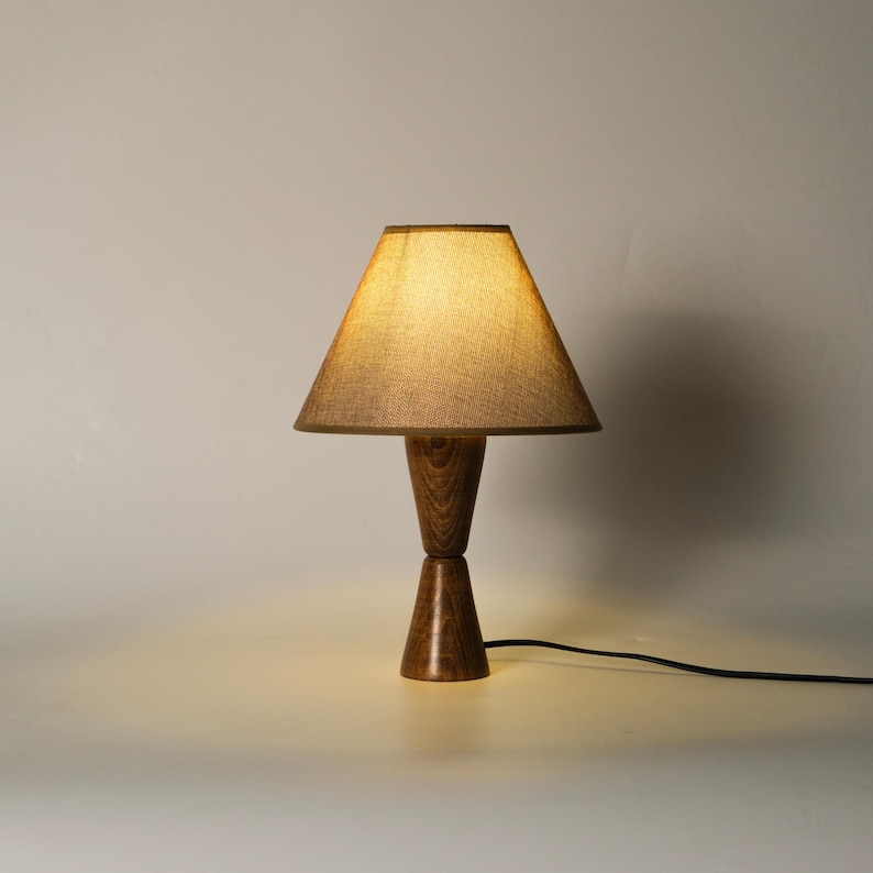 May include: A wooden table lamp with a conical lampshade. The lampshade is a light brown color, and the base is made of dark brown wood. The lamp has a classic design and a black power cord.