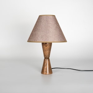 May include: A wooden table lamp with a brown lampshade. The lampshade is a light brown color with a tan trim. The base of the lamp is made of wood and has a unique hourglass shape. The lamp is plugged into a black electrical cord.