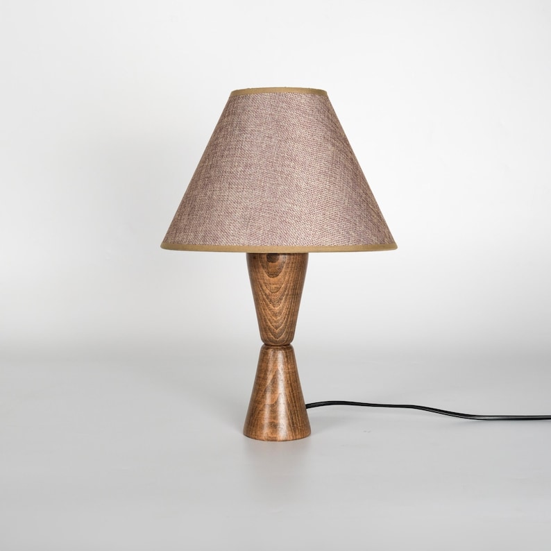 May include: A wooden table lamp with a brown lampshade. The lampshade is a light brown color with a tan trim. The base of the lamp is made of wood and has a unique hourglass shape. The lamp is plugged into a black electrical cord.
