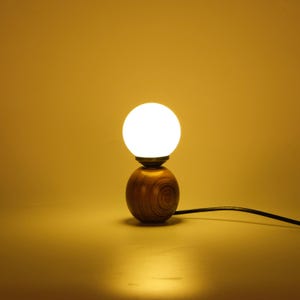 May include: A small wooden table lamp with a spherical, glowing white light bulb. The lamp has a round, wooden base and a black power cord. The lamp is on a yellow background.