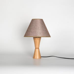 May include: A wooden table lamp with a light brown lampshade. The lampshade is cone-shaped with a tan trim. The base is made of light-colored wood and has a unique hourglass shape. A black power cord extends from the base.