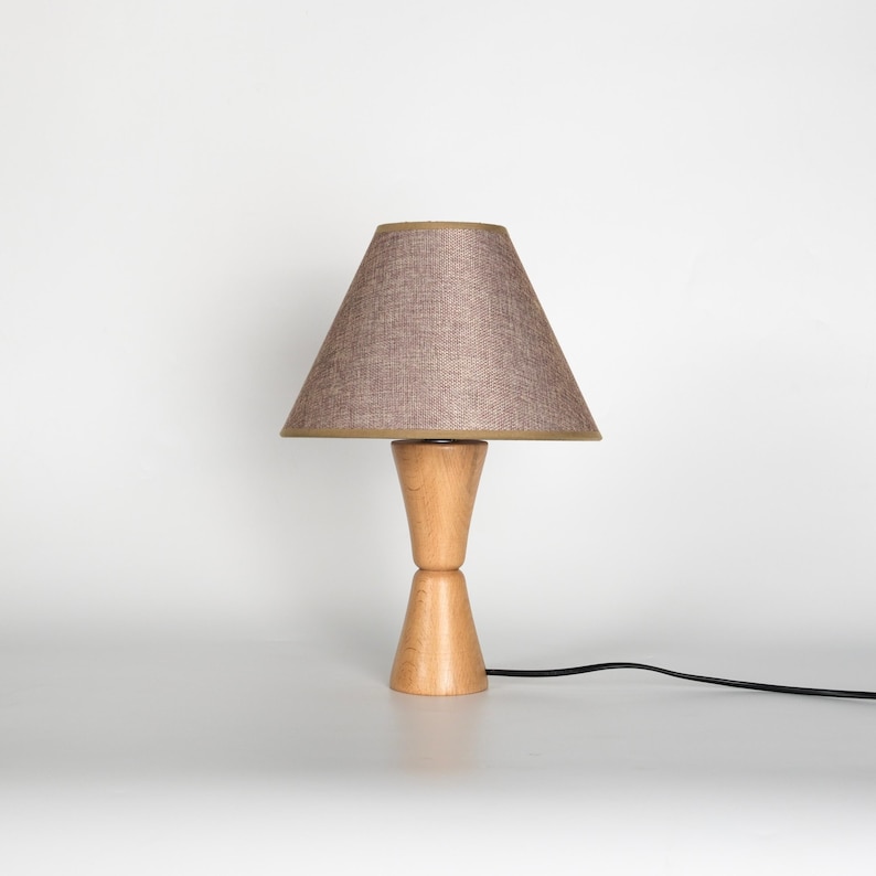 May include: A wooden table lamp with a light brown lampshade. The lampshade is cone-shaped with a tan trim. The base is made of light-colored wood and has a unique hourglass shape. A black power cord extends from the base.
