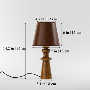 May include: A wooden table lamp with a brown fabric lampshade. The lamp is 14.2 inches tall and the shade is 4.7 inches in diameter. The lamp has a 7 inch tall base and a 3.1 inch wide base.