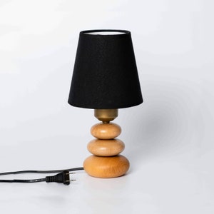 May include: A black lampshade with a white interior sits atop a wooden base made of three stacked circles. The lamp has a black cord and plug.
