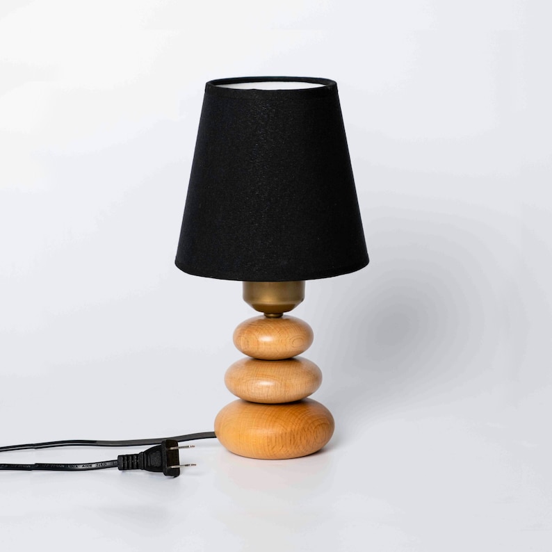May include: A black lampshade with a white interior sits atop a wooden base made of three stacked circles. The lamp has a black cord and plug.