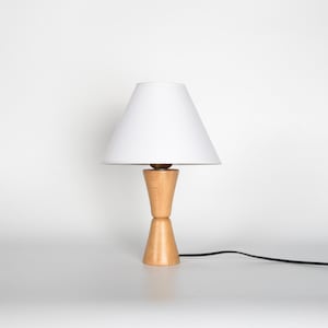 May include: A small, wooden table lamp with a white lampshade. The lamp has a light-colored, hourglass-shaped base and a brass-colored accent. A black power cord extends from the base.