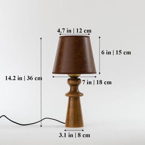 May include: A wooden table lamp with a brown fabric lampshade. The lamp is 14.2 inches tall and the shade is 4.7 inches in diameter. The lamp has a 7-inch tall base and a 3.1-inch wide base.