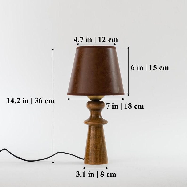 May include: A wooden table lamp with a brown fabric lampshade. The lamp is 14.2 inches tall and the shade is 4.7 inches in diameter. The lamp has a 7-inch tall base and a 3.1-inch wide base.