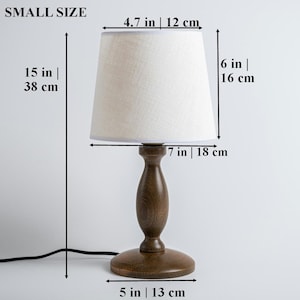 May include: A small, wooden table lamp with a white linen lampshade. The lamp is 15 inches tall and the shade is 6 inches tall. The base of the lamp is 5 inches in diameter.