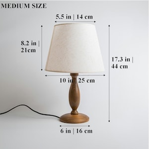 May include: A wooden table lamp with a beige fabric lampshade. The lamp is 17.3 inches tall, 10 inches wide, and has a 6-inch base. The lampshade is 5.5 inches wide and 8.2 inches tall.
